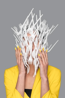 Woman face covered in plastic tableware standing next to a ultimate gray wall